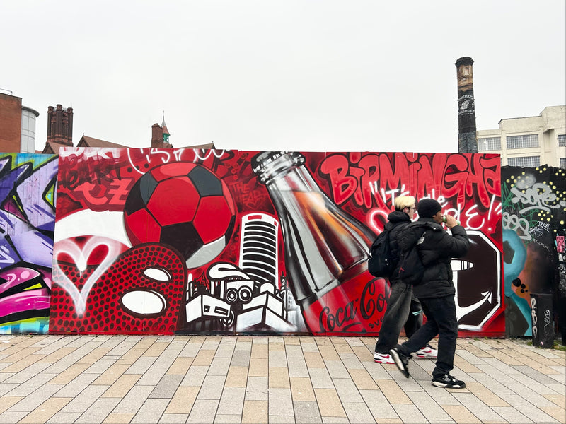 Two people walking past a large colorful mural with various graphics on a street.