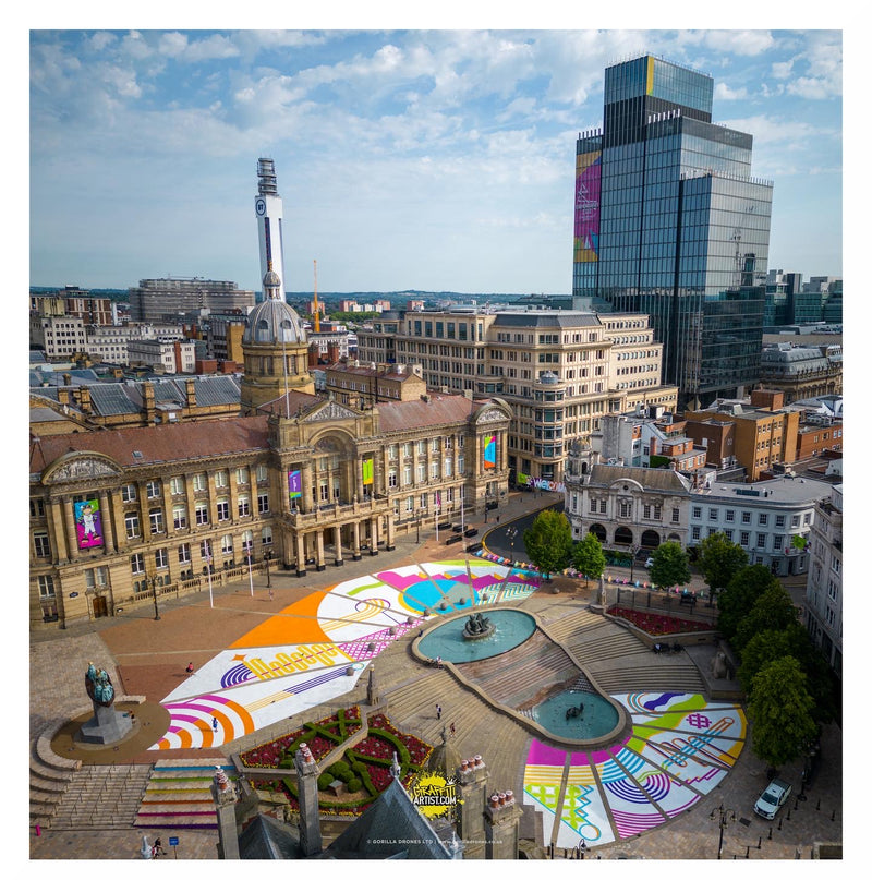 Aerial view of a city square with colorful artwork on the ground, surrounded by buildings.