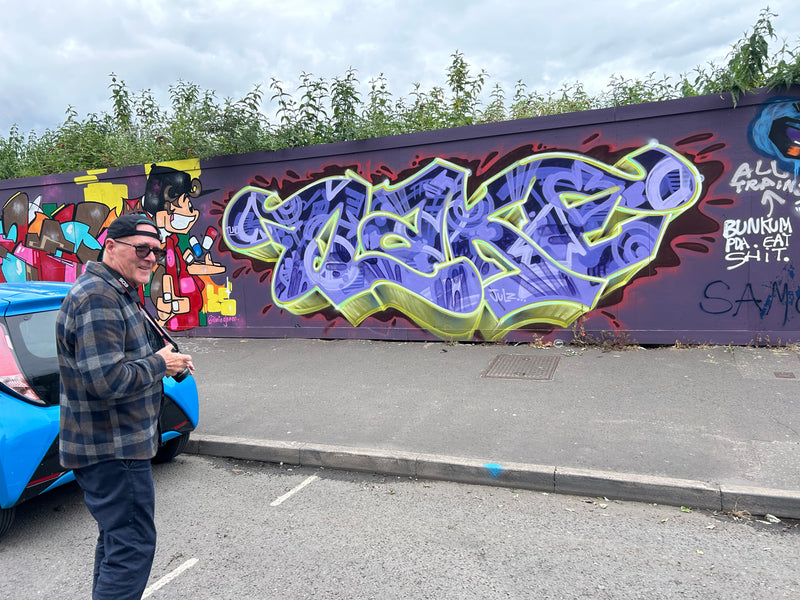 Man standing next to a blue car with graffiti art on a wall in the background
