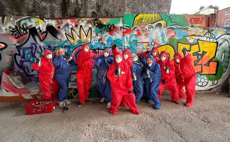 Group of people in colorful outfits standing in front of a graffiti-covered wall.