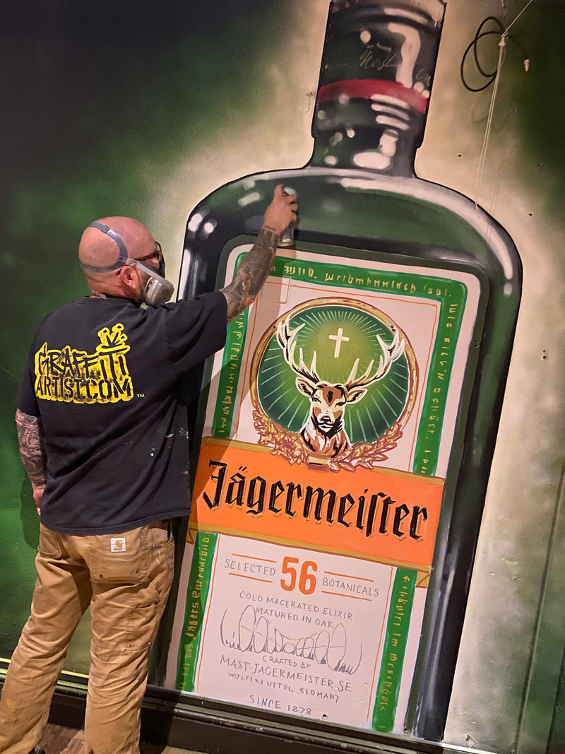 Person painting a large bottle with Jägermeister label on a green surface