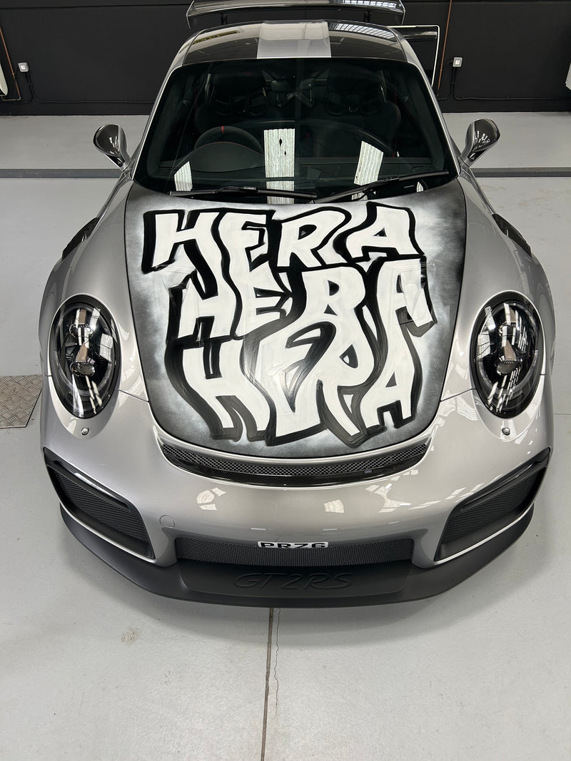 Car with a prominent Keith Haring-style graphic on its hood, parked indoors.