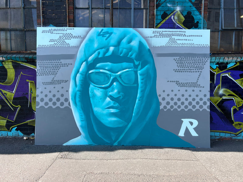 Blue hooded figure with goggles on a promotional board against a graffiti-covered wall.